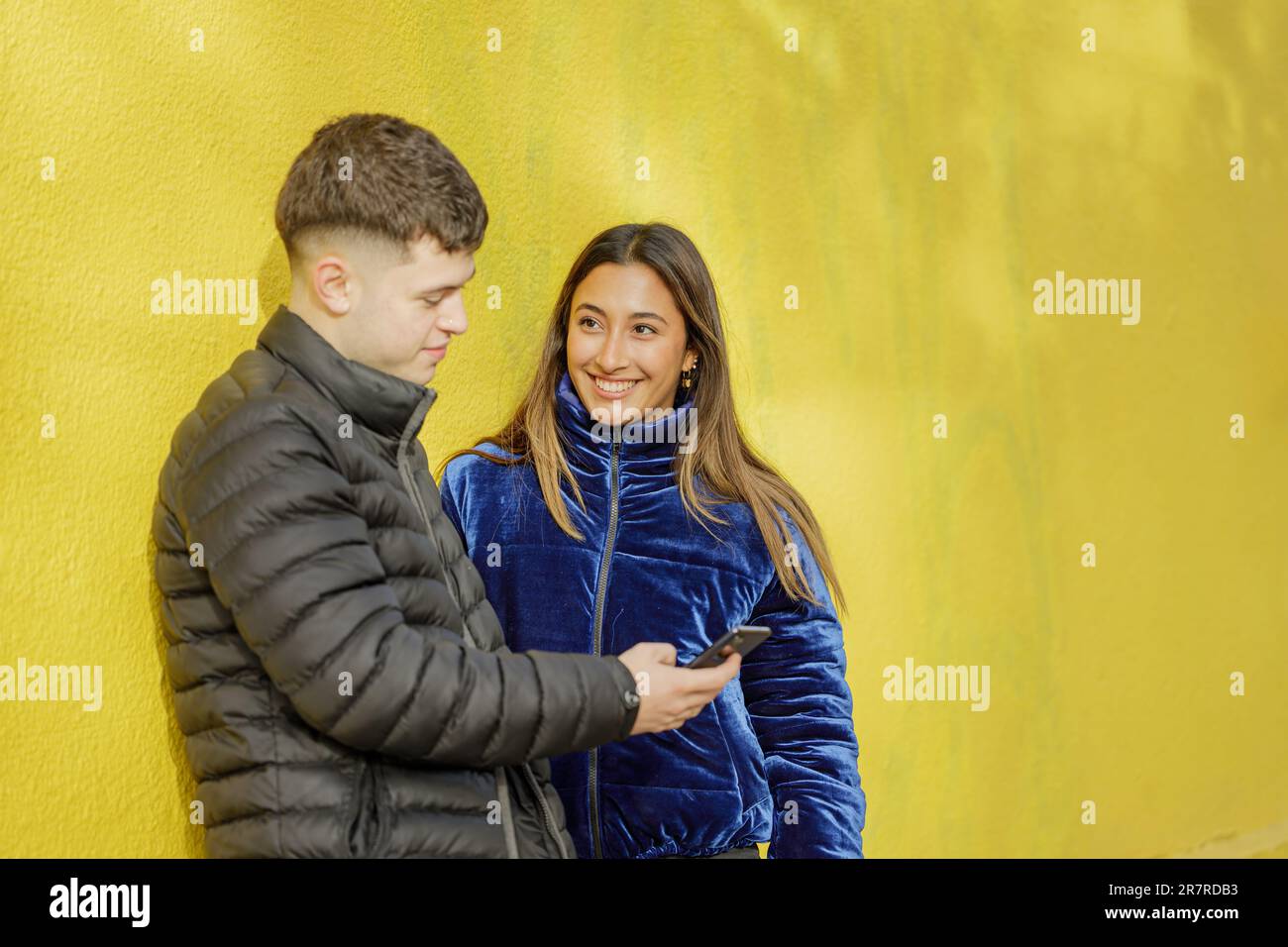 Couple of boy and girl leaning on a yellow wall watching the mobile phone with copy space. Stock Photo