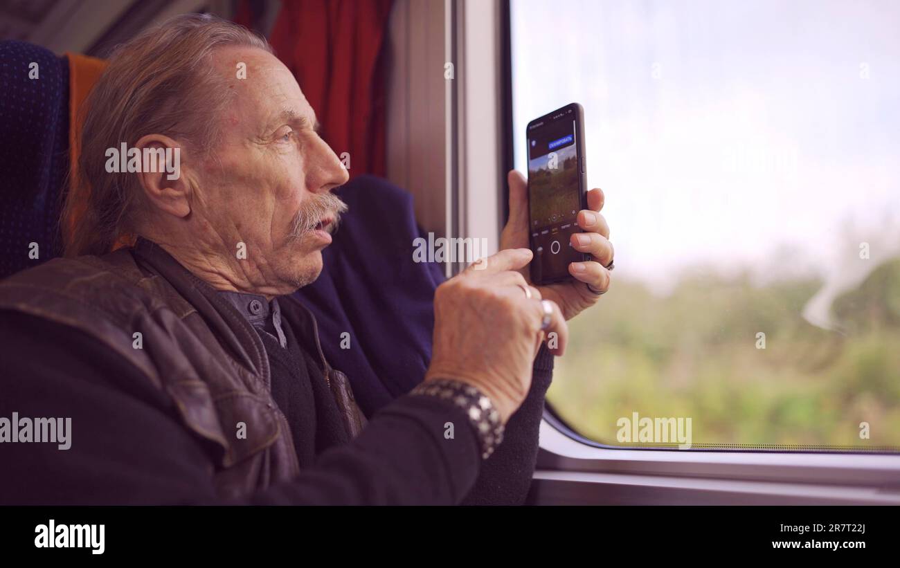 Elderly man traveling by train and taking pictures of the landscape through the window using a smartphone Stock Photo