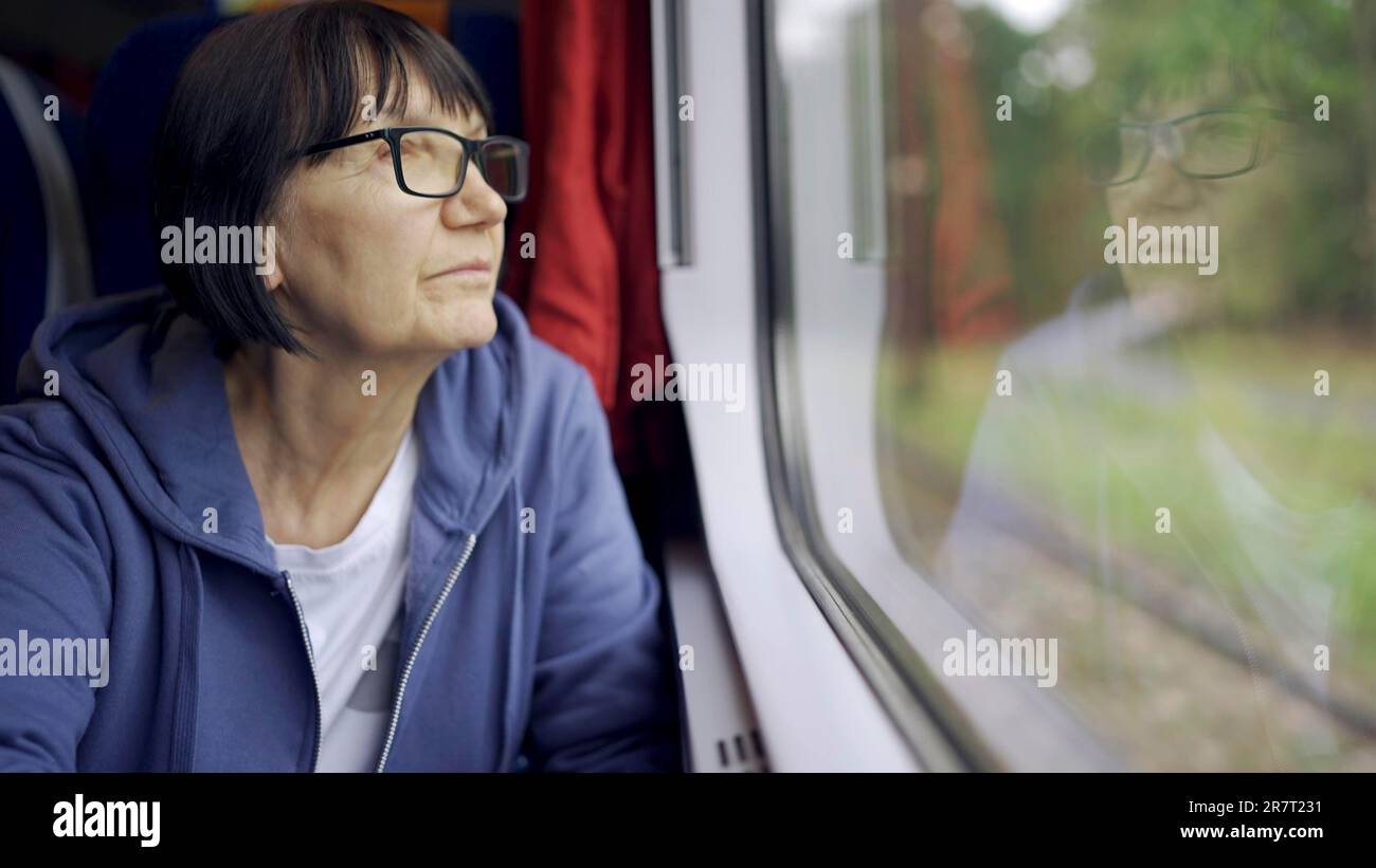 Elderly lady in glasses travels in train and looking out the window reflected in the glass Stock Photo