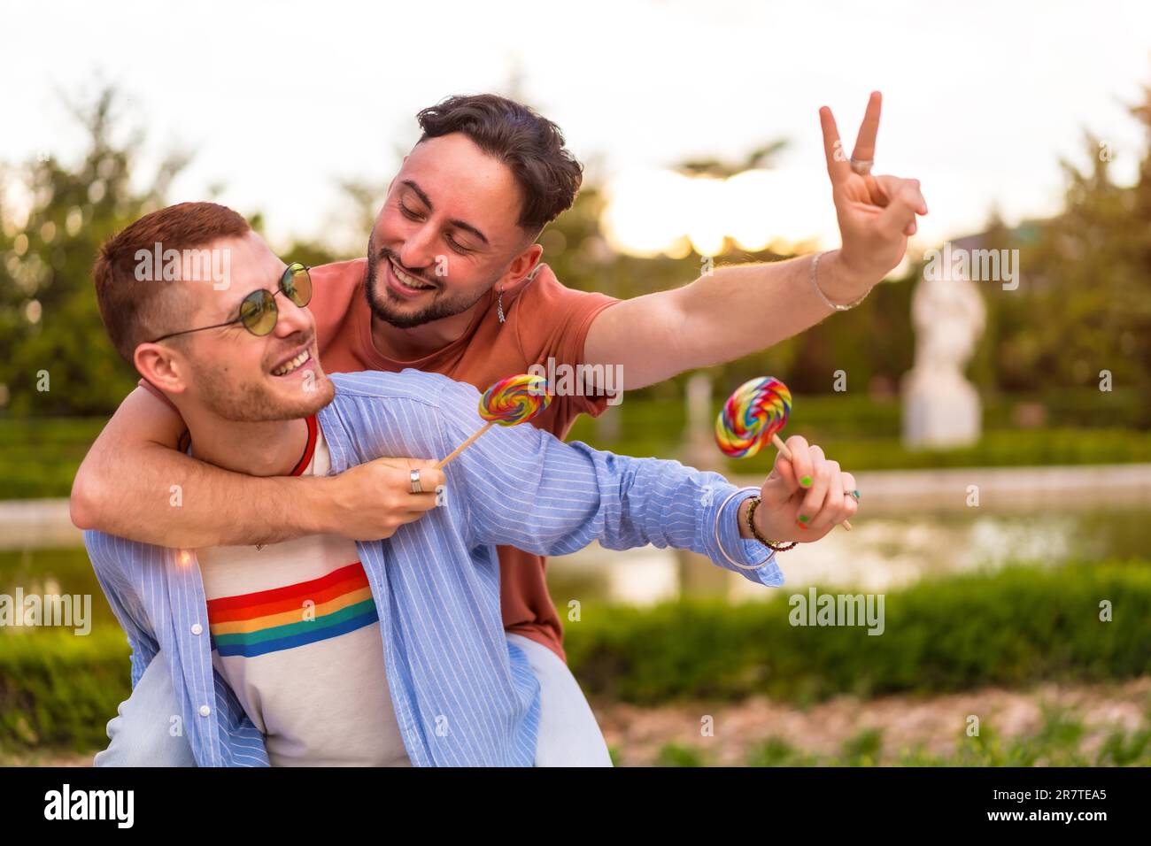 Portrait of gay boyfriend playing and one climbed on his back running eating a lollipop in the park on sunset in the city. lgbt concept Stock Photo