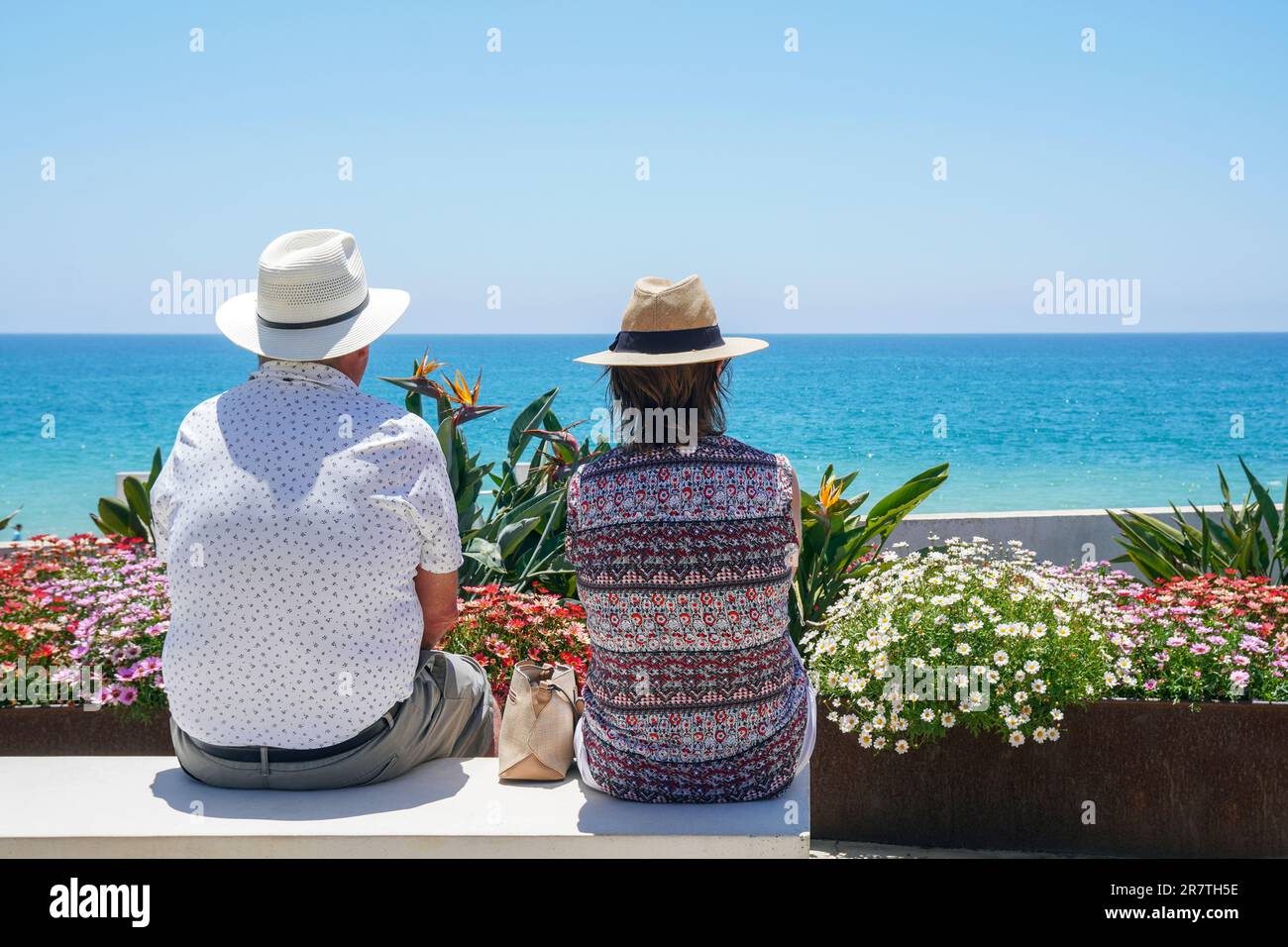 Man and woman sitting on a public bench and looking over the small garden and beach to the sea and horizon, Quinta do Lobo, Algarve, Portugal Stock Photo