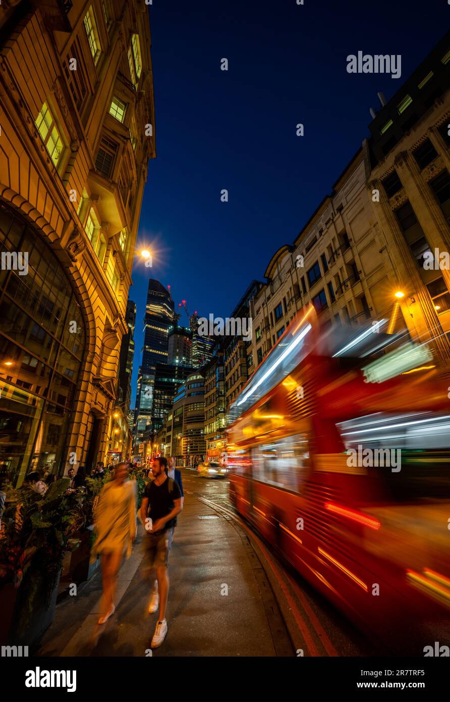 London, UK: Gracechurch Street in the City of London at night. People walk along the pavement as a red London bus passes by. Tall buildings behind. Stock Photo