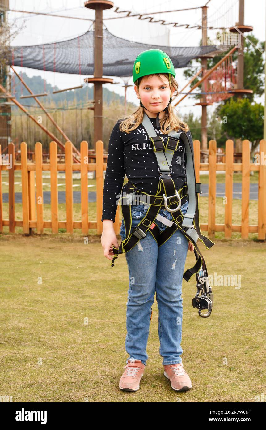 Teenage teen girl in climbing harness equipment, green sports safety helmet. Rope amusement park. Fastening attaching carabiner to safety rope. Hangin Stock Photo