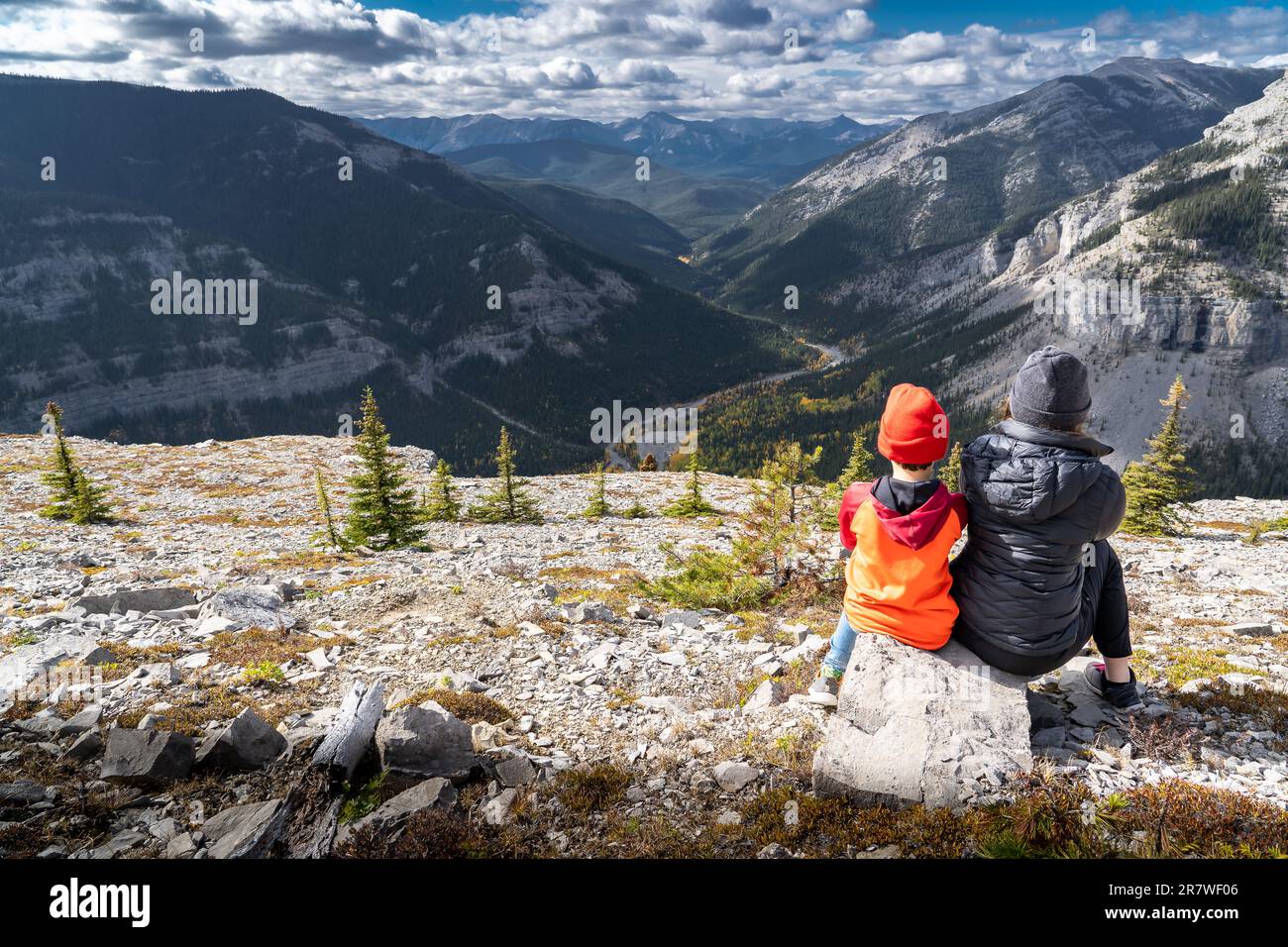 Brother and sister sitting on a rock overlooking the Canadian Rocky Mountains with valley trees in fall colours on Moose Mountain neat Bragg Creek Alb Stock Photo