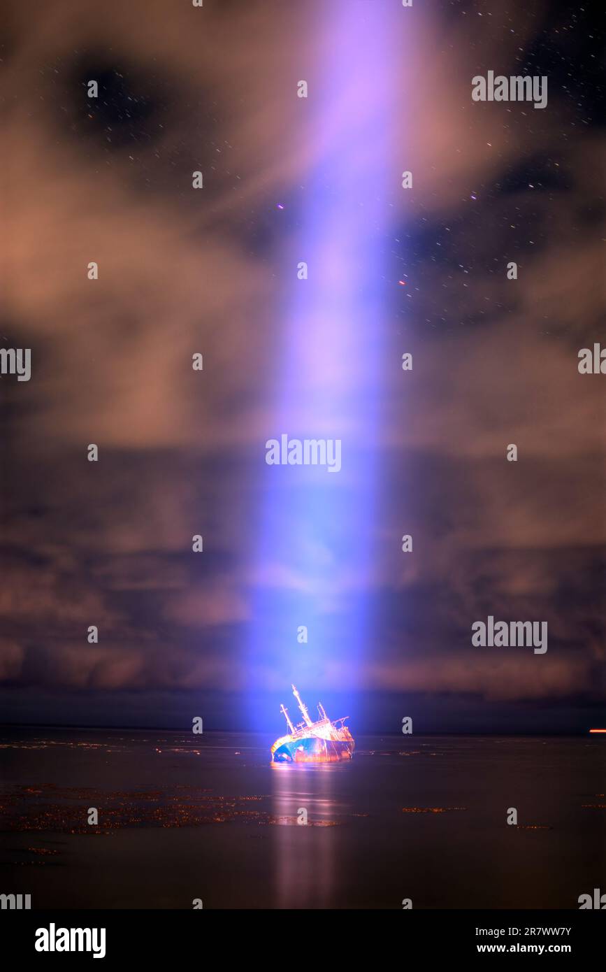 A bright, focused beam of light is directed from the sky at a ship that has run aground and lists on its side Stock Photo