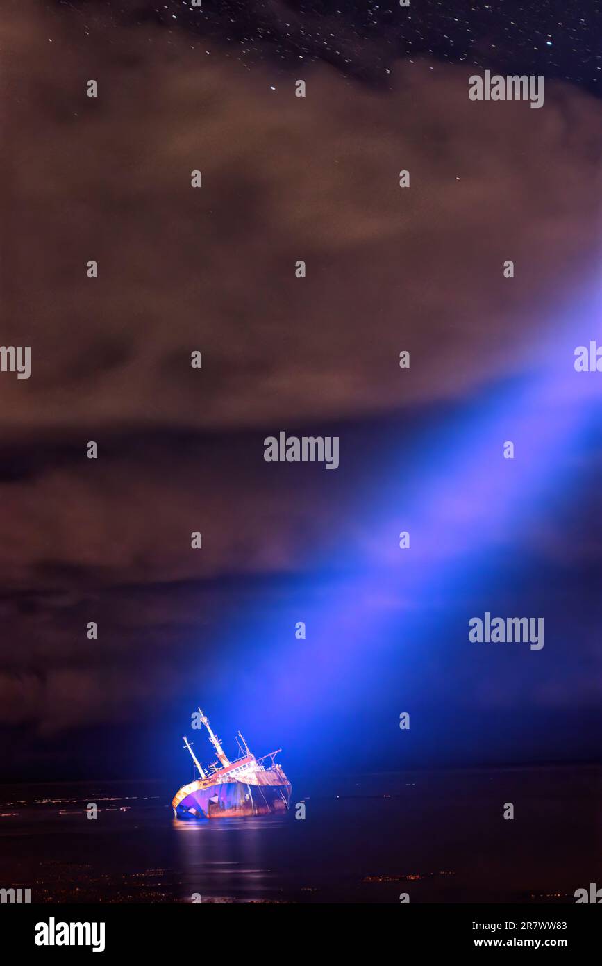 A bright, focused beam of light is directed from the sky at a ship that has run aground and lists on its side Stock Photo