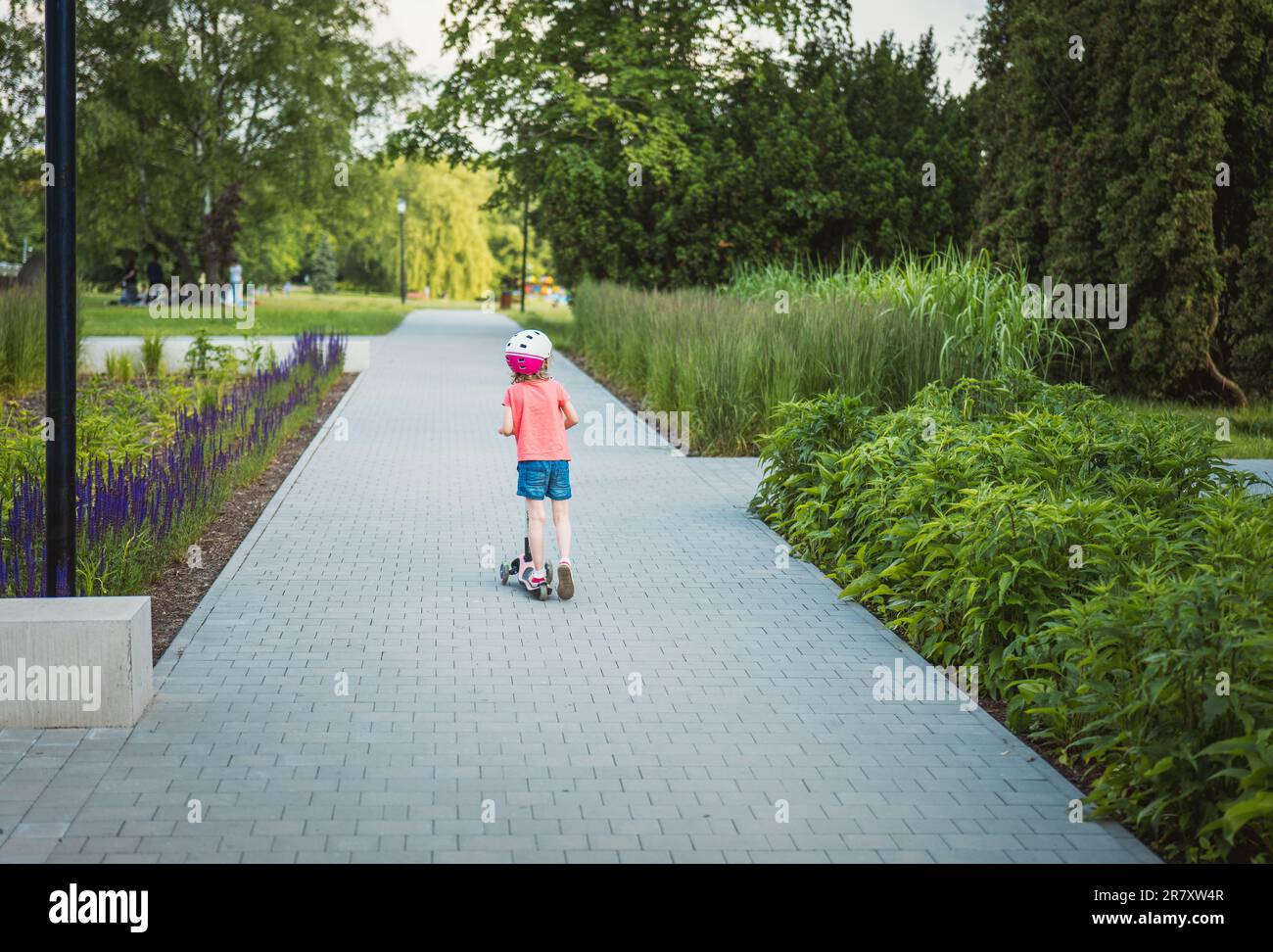 A little girl rides a scooter in a beautiful park full of greenery. Stock Photo