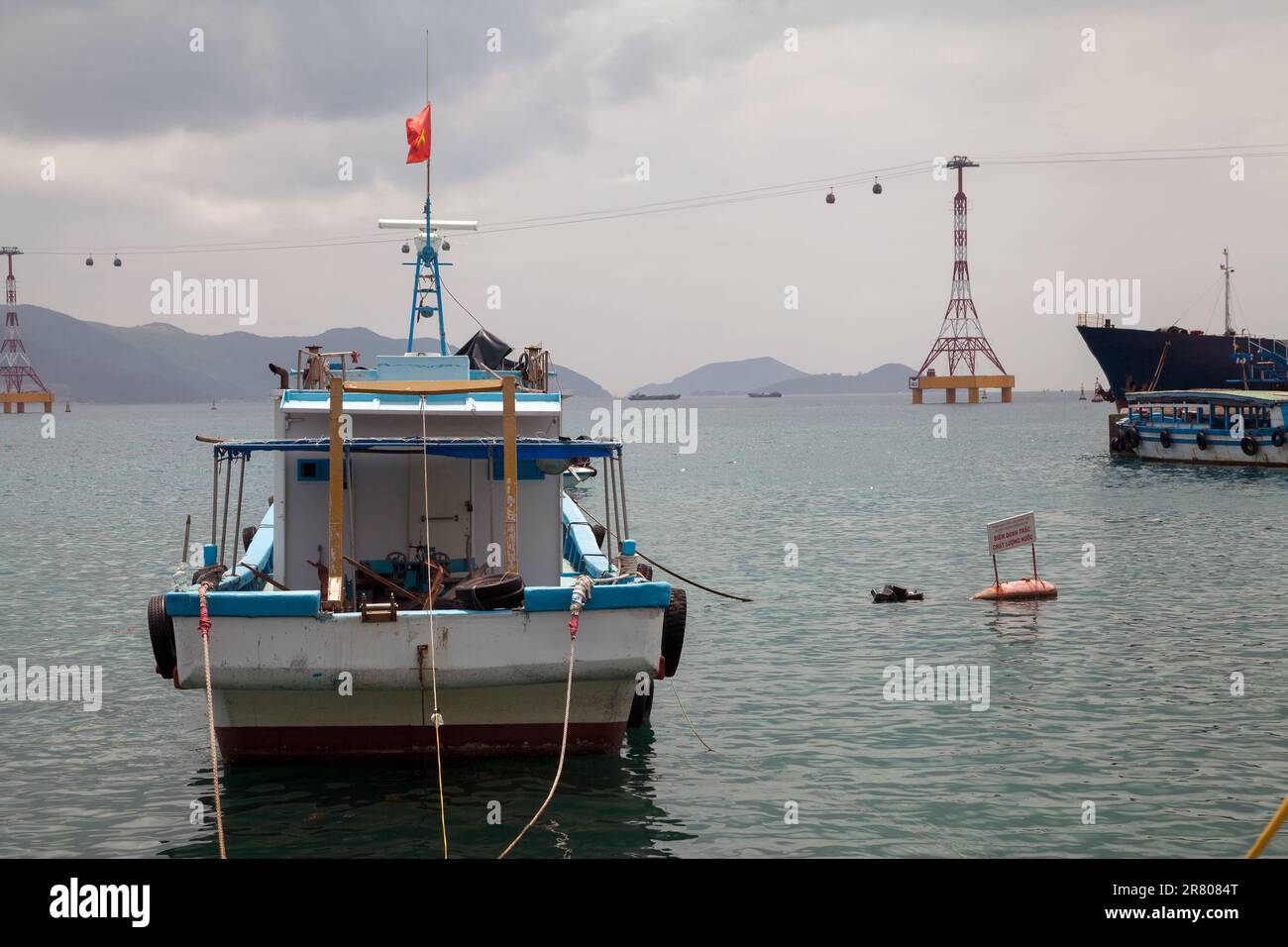 Rear view of an old boat in a harbor against a beautiful scenery with mountains and a cableway. Sea bay with boats and a cable car in Nha Trang, Vietn - Stock Image