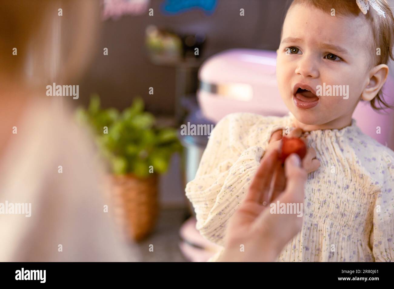 Mom feeding her baby girl with sour strawberries in the kitchen. Rejecting food Stock Photo