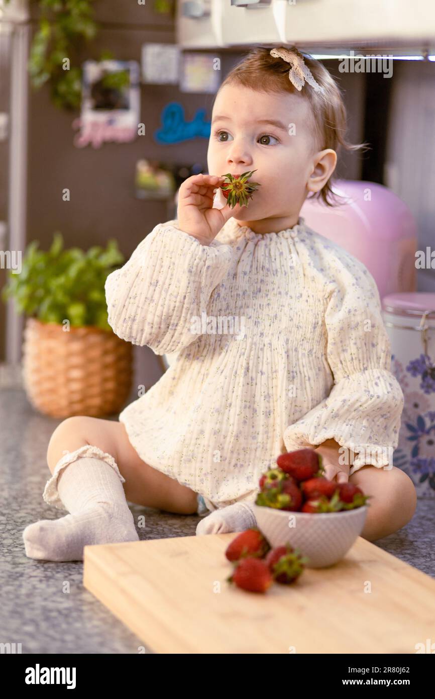 Little baby girl sitting on the kitchen table by herself, eating strawberries Stock Photo