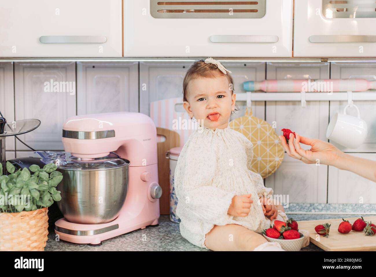 Mom feeding her baby girl with sour strawberries in the kitchen. Rejecting food Stock Photo