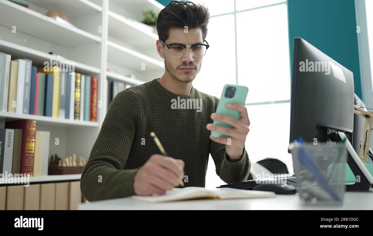 Young hispanic man student using smartphone writing on notebook at ...