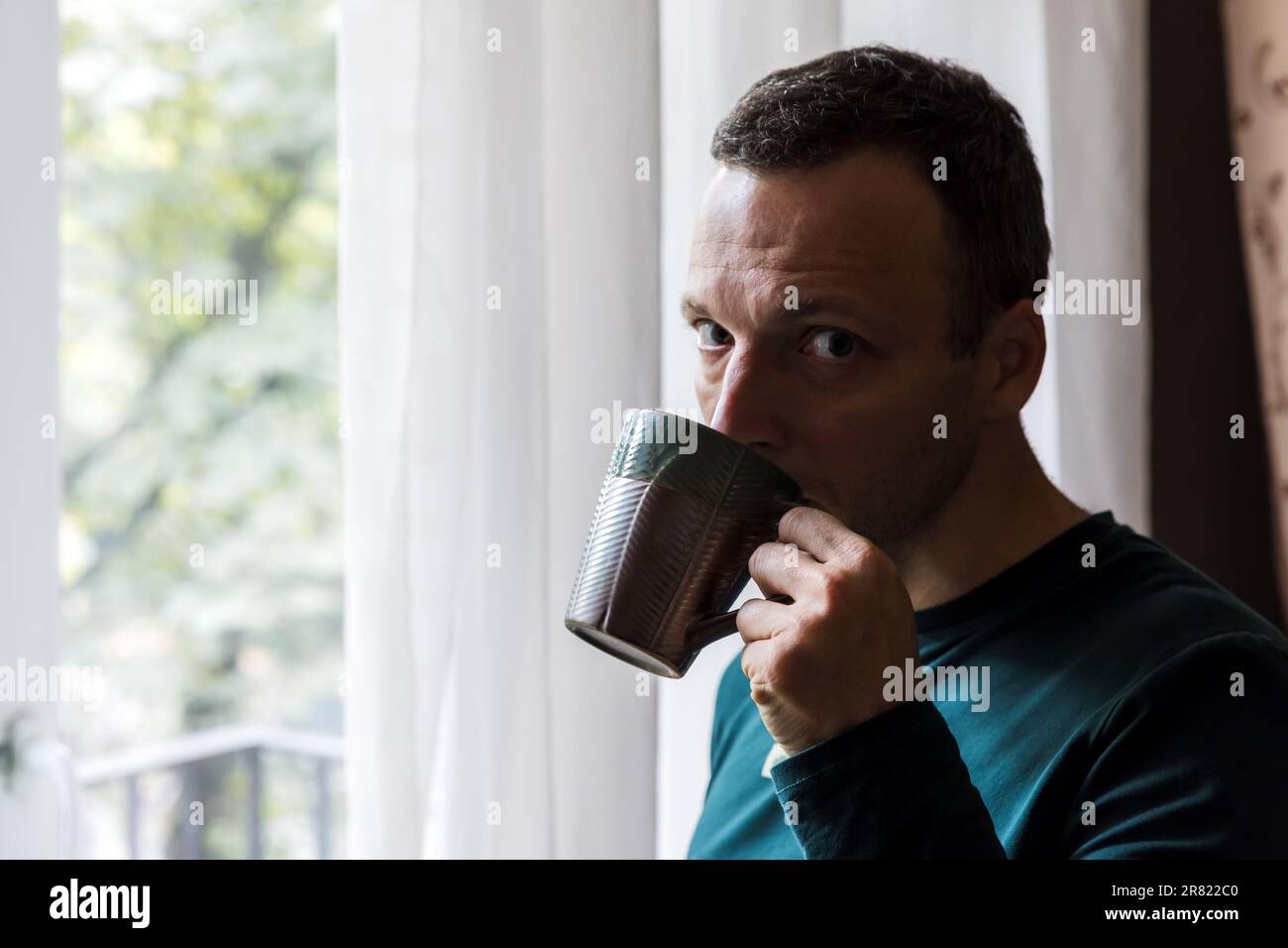 Young adult Caucasian man drinks coffee near a window on a daytime Stock Photo