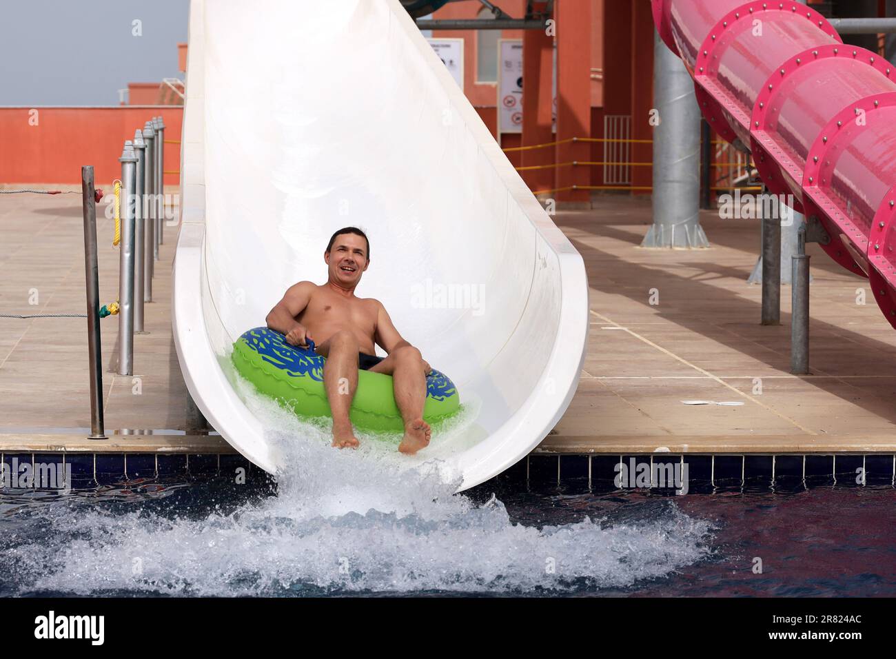Happy man in inflatable circle rides down on water slide at aqua park. Riding on water tube, enjoying summer holiday, beach resort Stock Photo