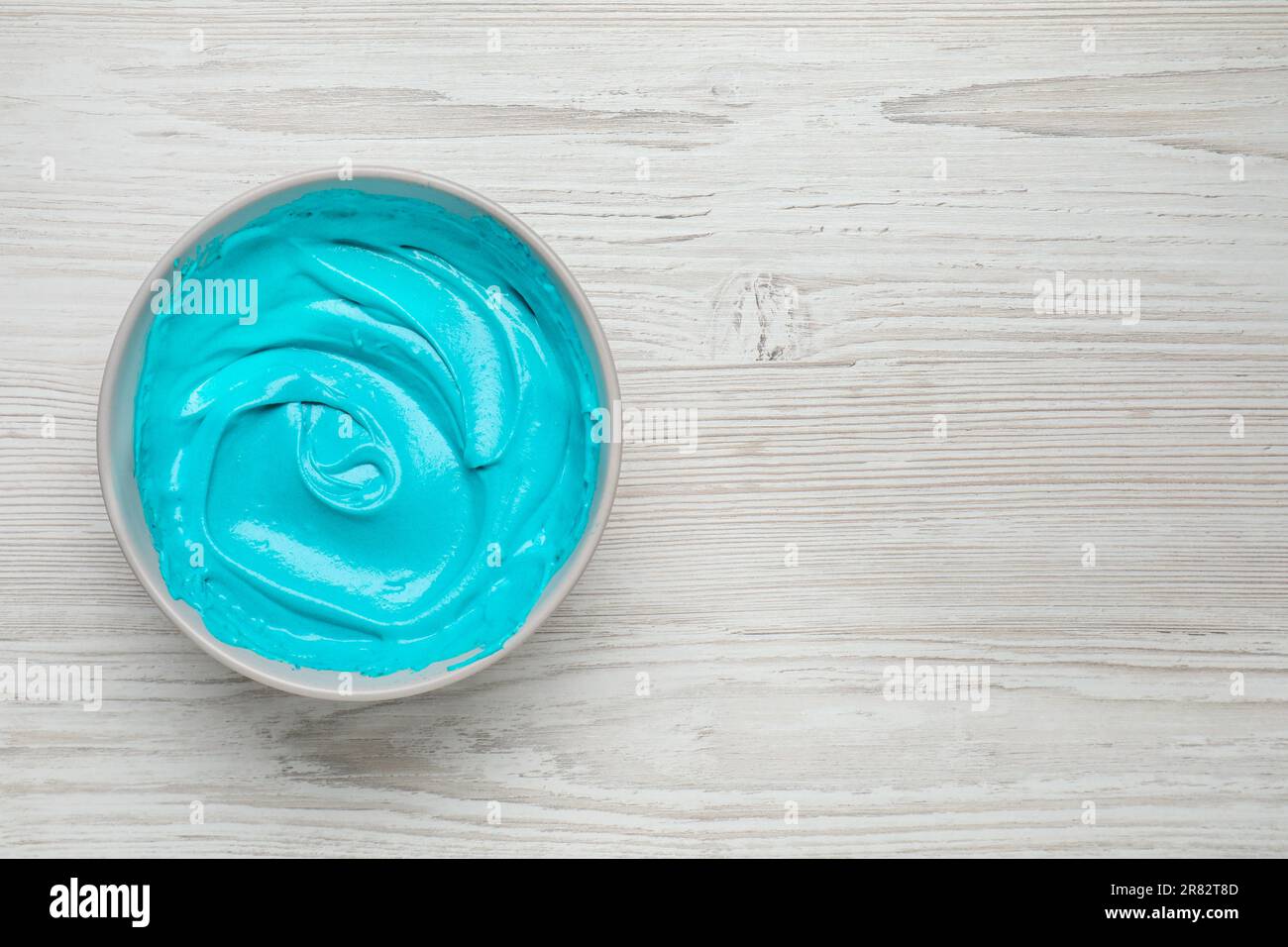 Bowl of cream with light blue food coloring on white wooden table, top