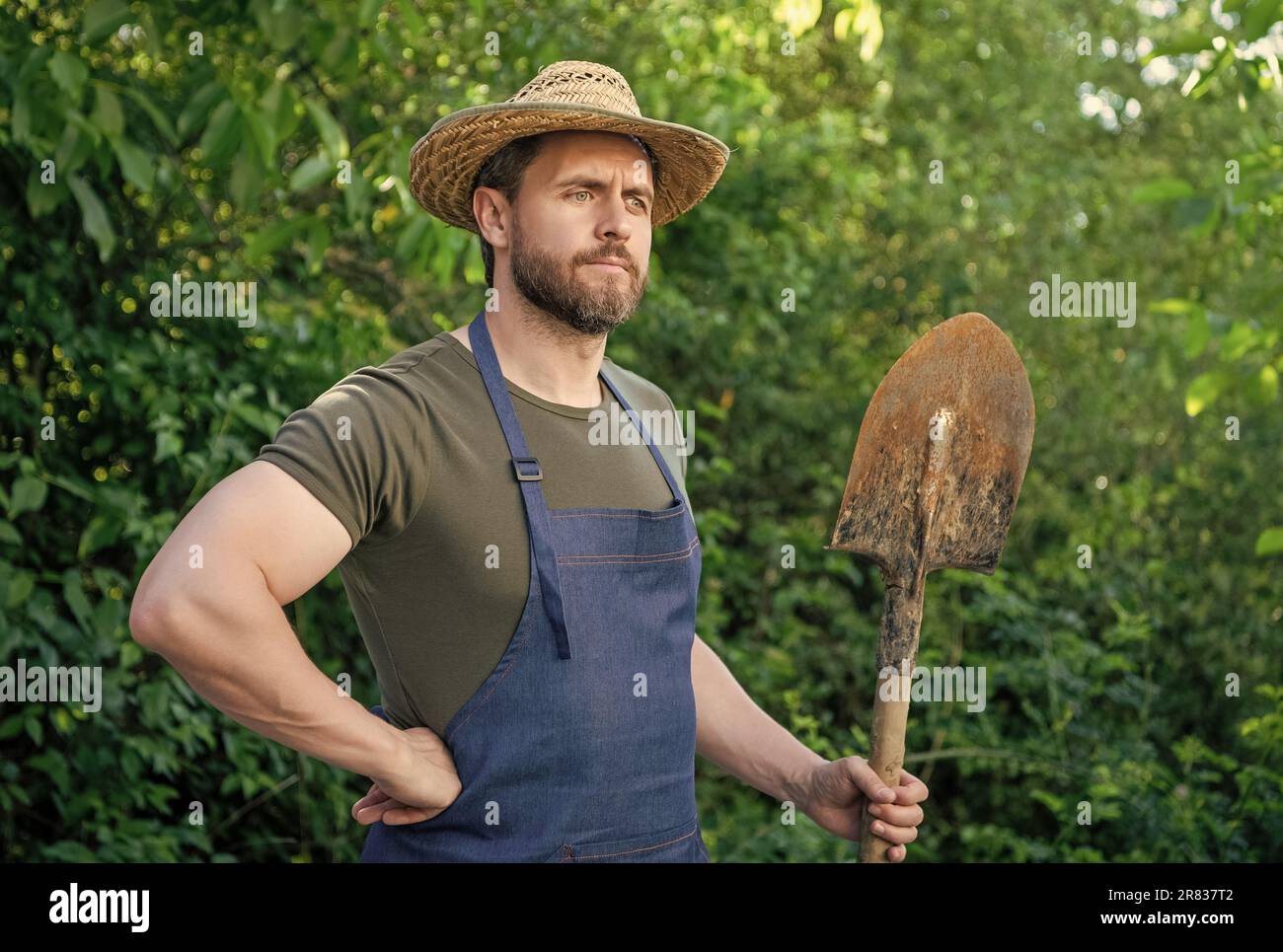 Thoughtful farmer man in farmers hat and gardening apron holding garden shovel Stock Photo