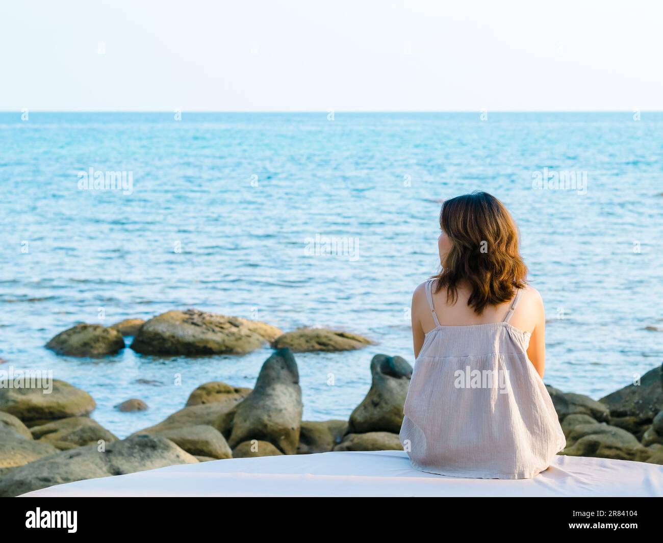 Back of Asian woman in casual dress sit in front of the beach and rock coast in summer. Lonely female relaxing at the sea and looking far away, seasca Stock Photo