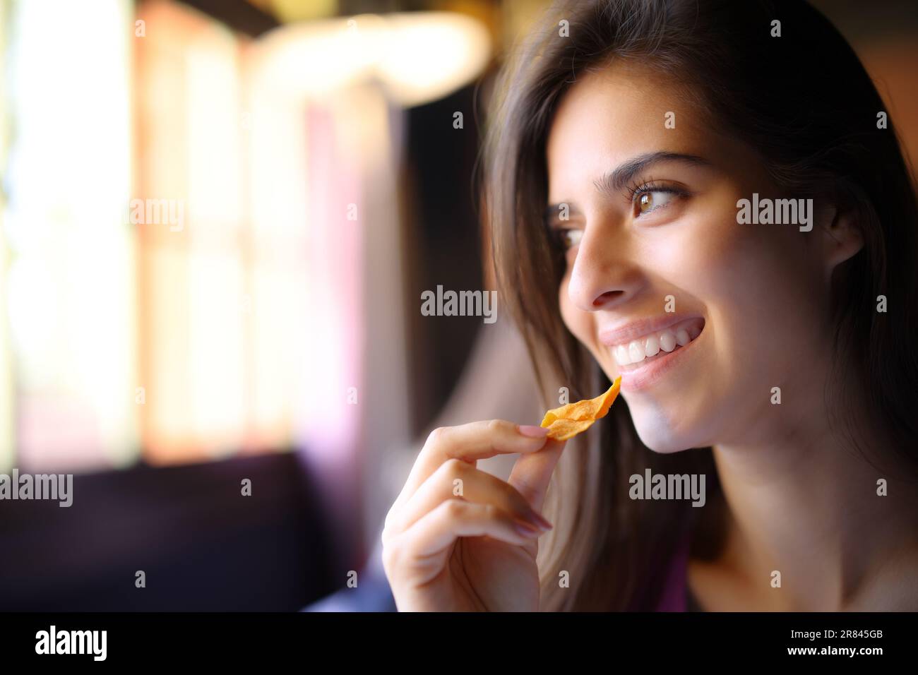 Happy restaurant customer eating chips looks at side through a window Stock Photo