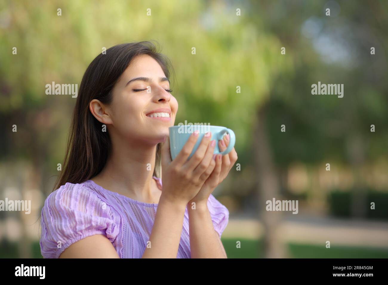 Happy woman holds coffee cup and smell it standing in a park Stock Photo