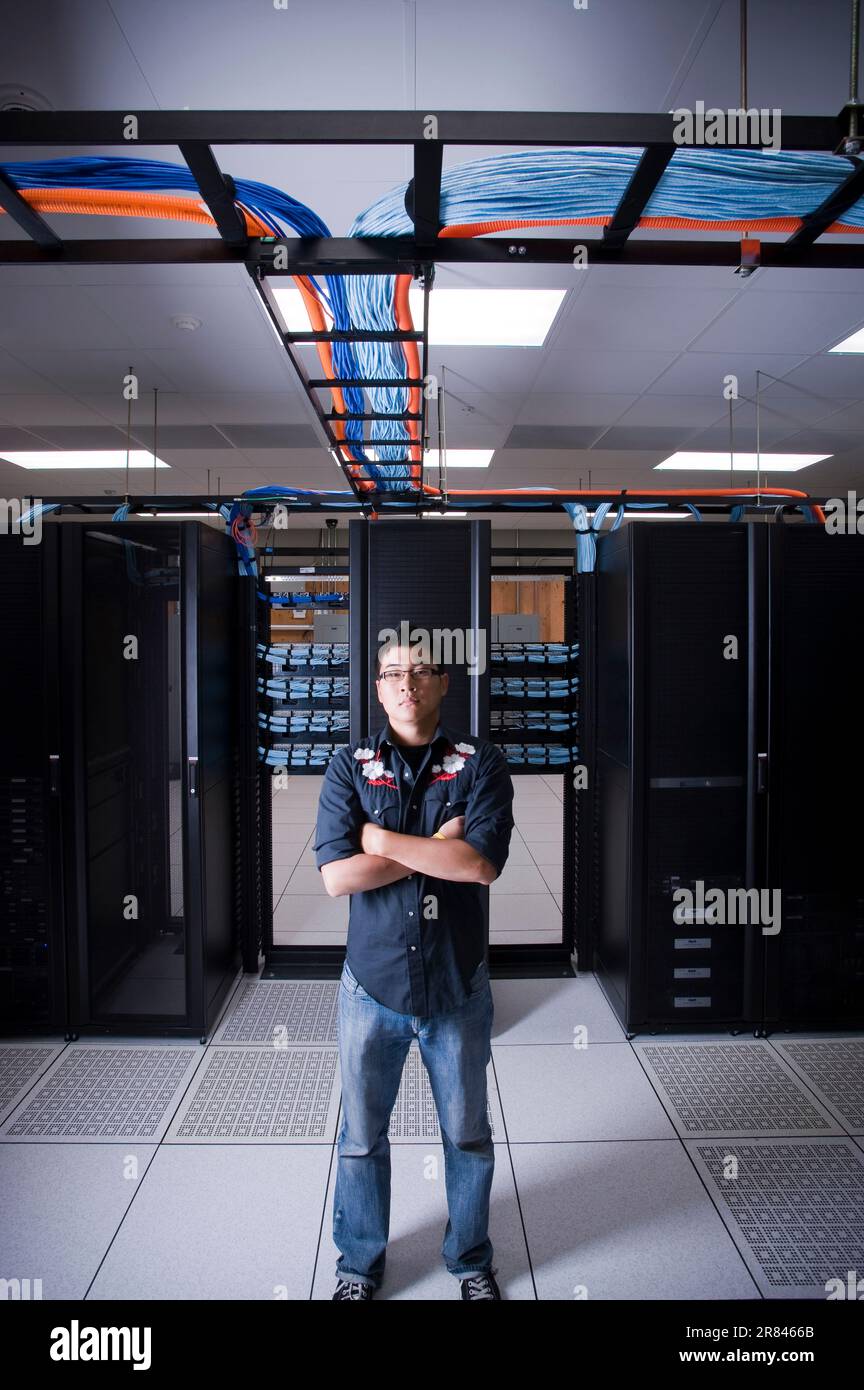 A young adult Asian male stands confidently in front of a row of new network servers in a server room. Stock Photo