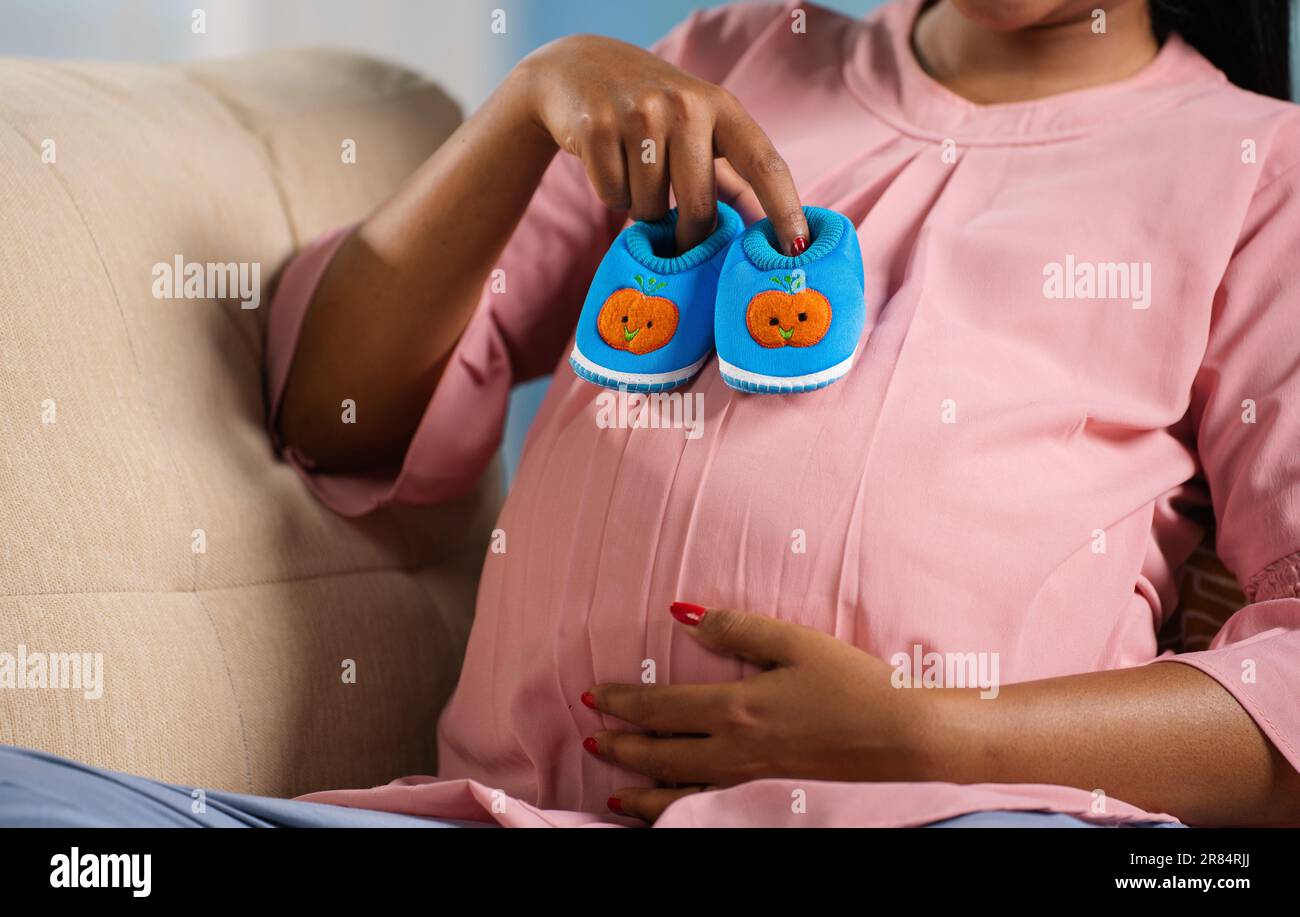 Close up headless shot of Indian pregnant woman playin by placing baby booties on tummy at home - concept of orenatal connection, motherhood and Stock Photo