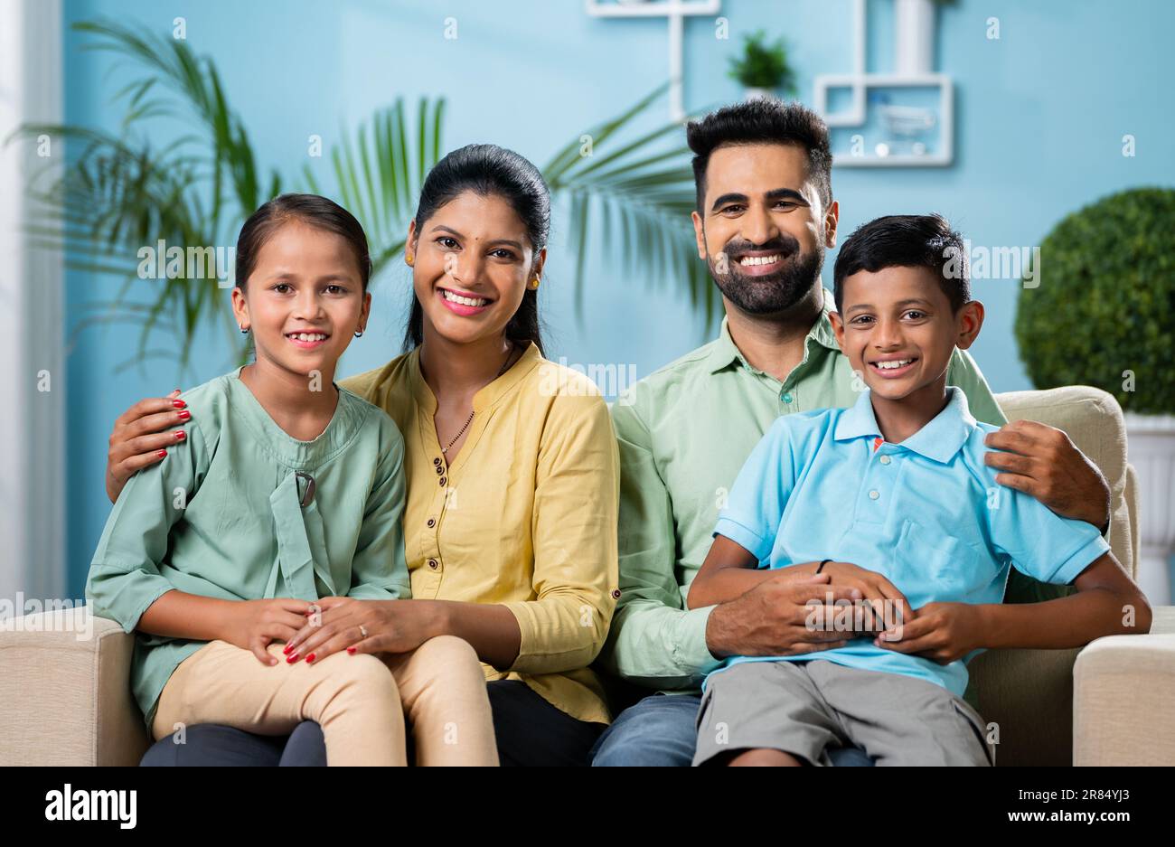Happy smiling indian couple with sibling kids looking or posing to camera while sitting on sofa at home - concept of nuclaer family, parenthood and Stock Photo