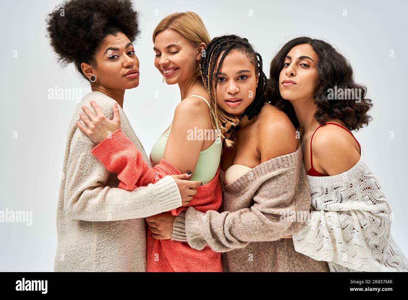 Portrait of group of multiethnic girlfriends in warm knitted jumpers hugging and standing together isolated on grey, different body types and self-acc Stock Photo