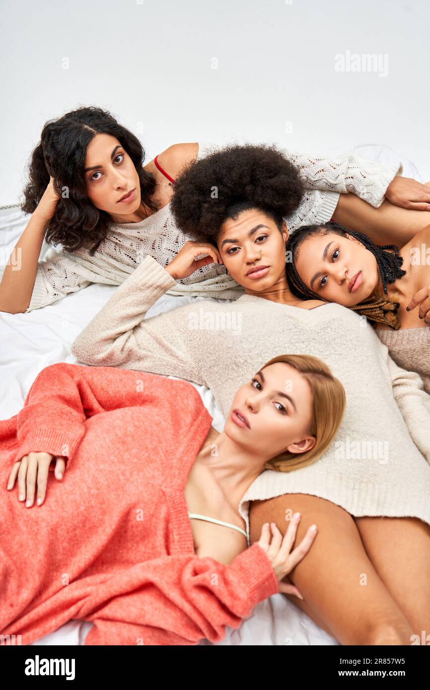 High angle view of multiethnic women in cozy knitted jumpers looking at camera while lying on bed isolated on grey, different body types and self-acce Stock Photo
