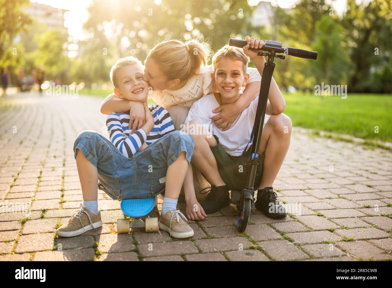 Joyful mother having fun with her two sons in park on sunny day. They are ridding skateboard and scooter. Stock Photo