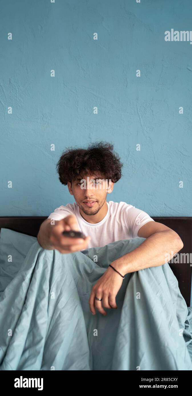 Young handsome man watches TV while sitting in bed in the morning, uses the remote control Stock Photo