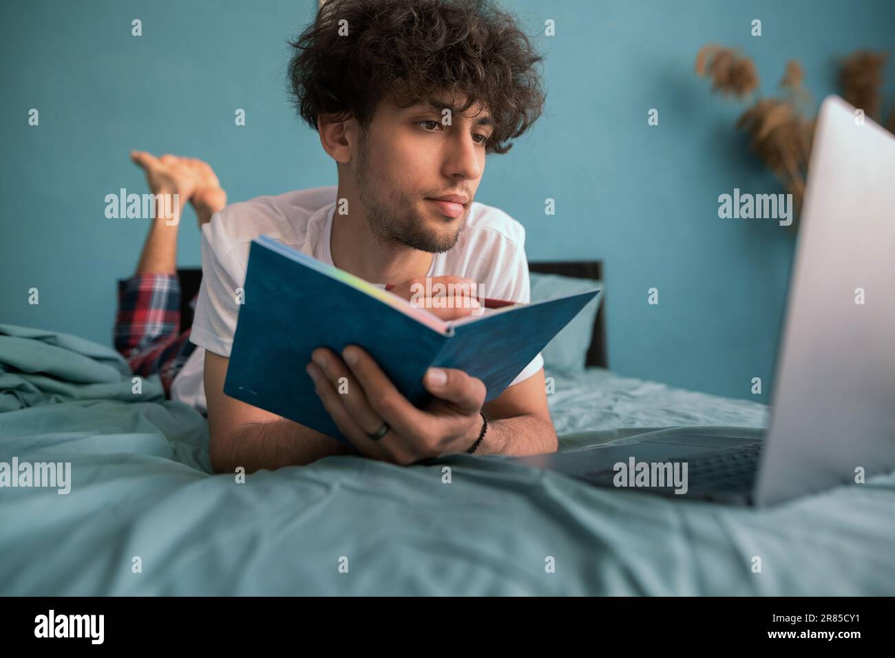 Curly young eastern guy studying in bed, using laptop, watching course on Internet or webinar, taking notes, copy space Stock Photo