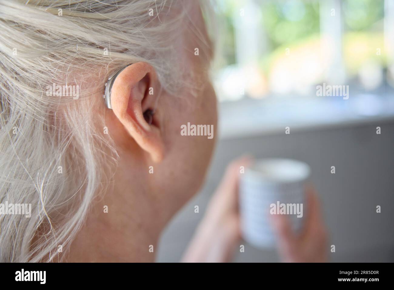 Close Up Of Senior Woman Wearing Behind The Ear Hearing Device Or Aid With Hot Drink At Home Stock Photo