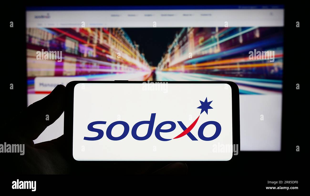 Person holding smartphone with logo of French facility management company Sodexo S.A. on screen in front of website. Focus on phone display. Stock Photo