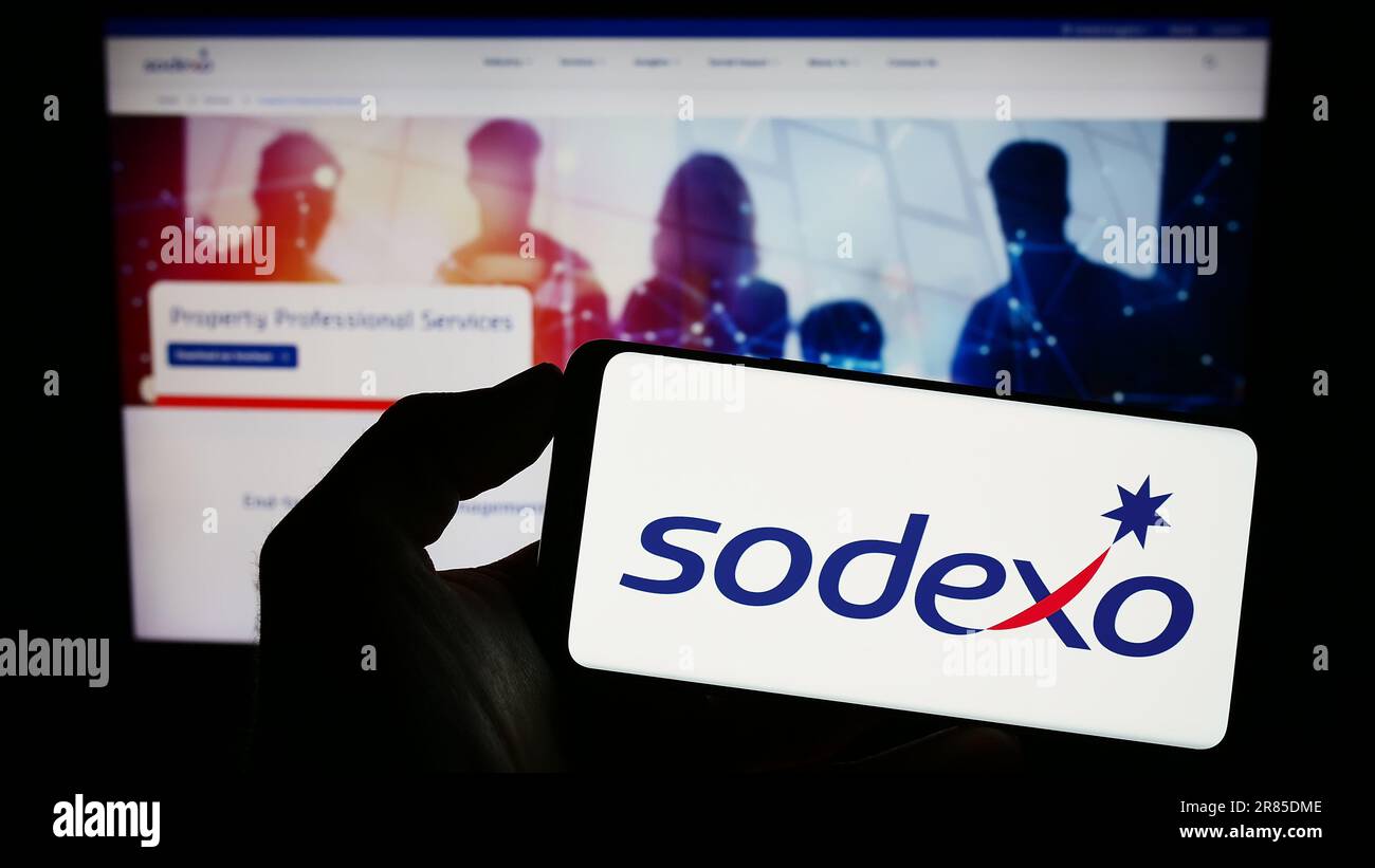 Person holding cellphone with logo of French facility management company Sodexo S.A. on screen in front of webpage. Focus on phone display. Stock Photo