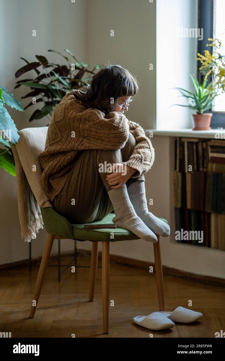 Lonely depressed teen girl hugging knees sitting alone on chair at home ...