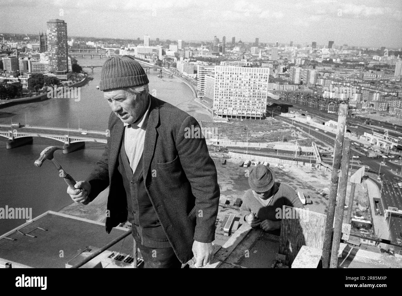 London Skyline 1970s. Irish Navvie Navvy an Irish labourer, a construction worker holding hammer at the top of and working on a Nine Elms tower block. London cityscape view, the  River Thames, Vauxhall Cross below and Waterloo in the distance. London, England circa 1974. HOMER SYKES Stock Photo
