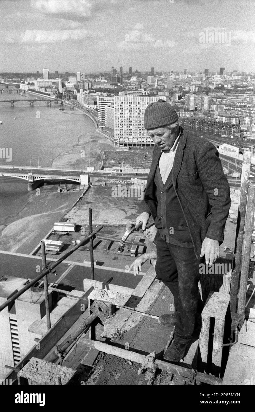 London Skyline 1970s. Irish Navvie Navvy an Irish labourer, a construction worker holding hammer at the top of and working on a Nine Elms tower block. London cityscape view, the  River Thames, Vauxhall Cross below and Waterloo in the distance. London, England circa 1974. HOMER SYKES Stock Photo