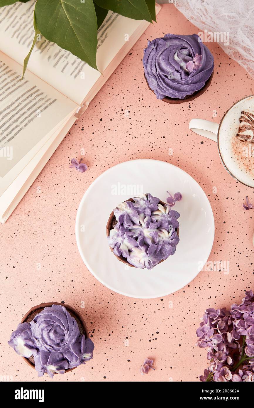 Aesthetic female lifestyle flat lay. Book and purple floral cake. Violet sweet no sugar dessert and coffee among lilac flowers. Stock Photo
