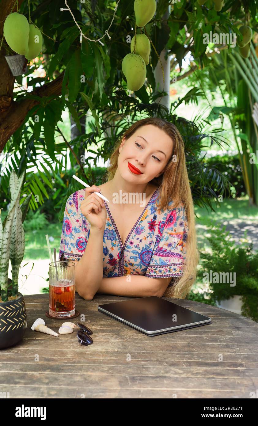 Beautiful blonde woman artist and illustrator thinking about a concept of her drawing, sitting by the table with electronical tablet and stylus Stock Photo