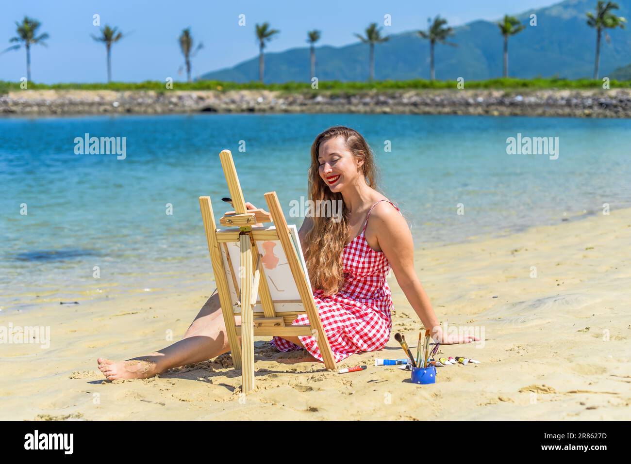 Young artist woman sitting at the beach with a jar with paint brushes ...