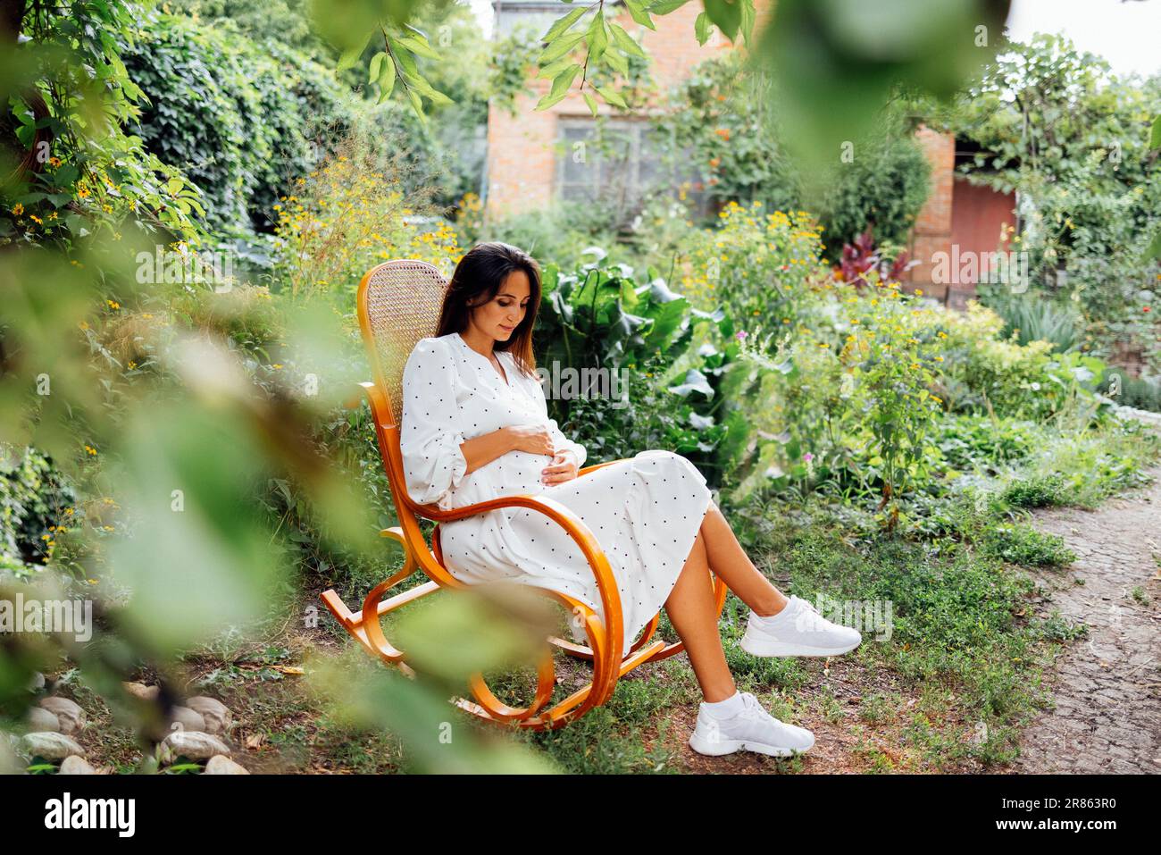 Charming pregnant brunette in elegant white dress is sitting on yellow rocking chair outdoors in garden. Young woman expecting baby holds her tummy an Stock Photo