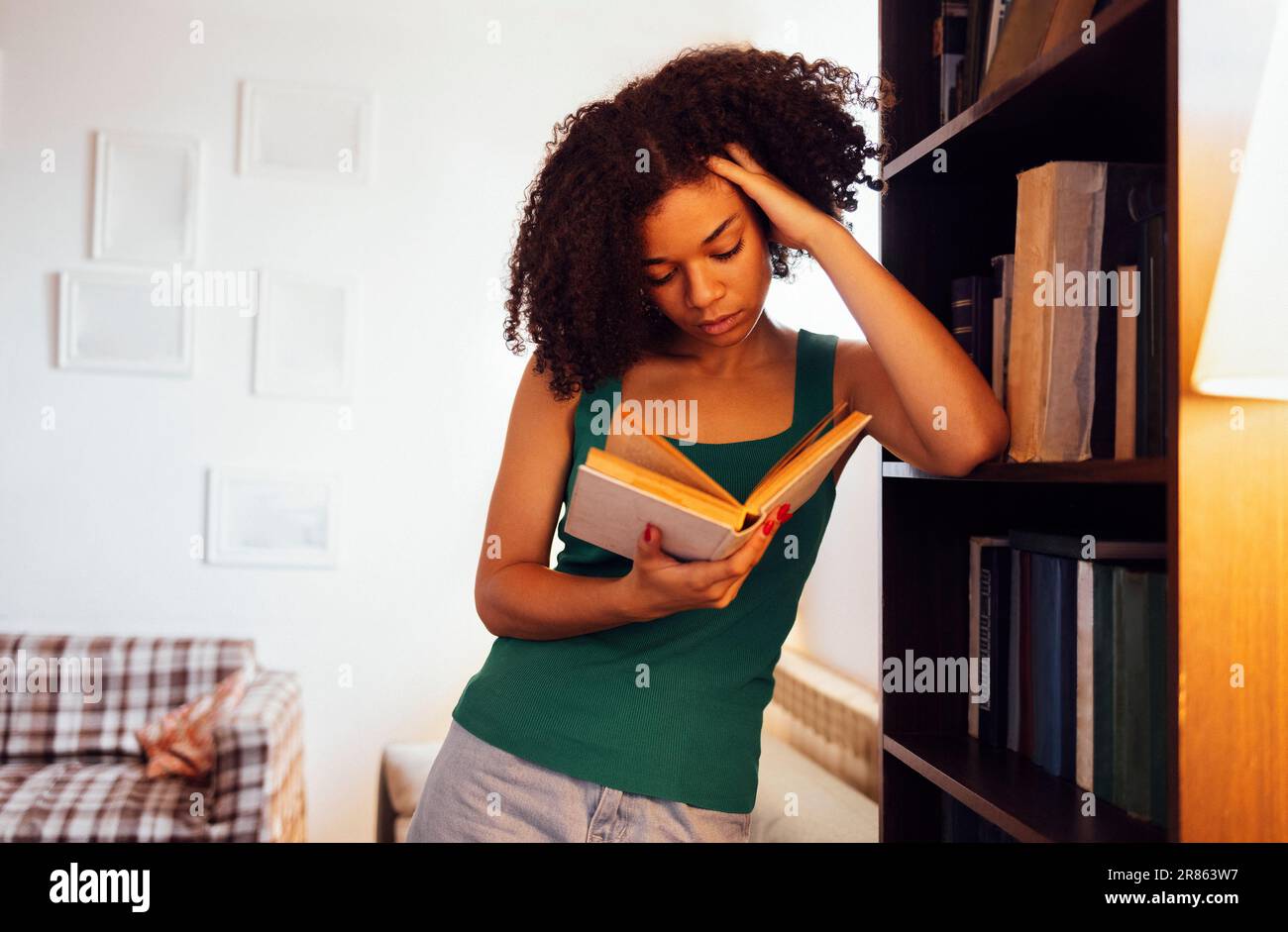 Thoughtful girl of mixed race holds and reads a book leaning on bookcase in cozy living room. Charming african female student studying literature at h Stock Photo