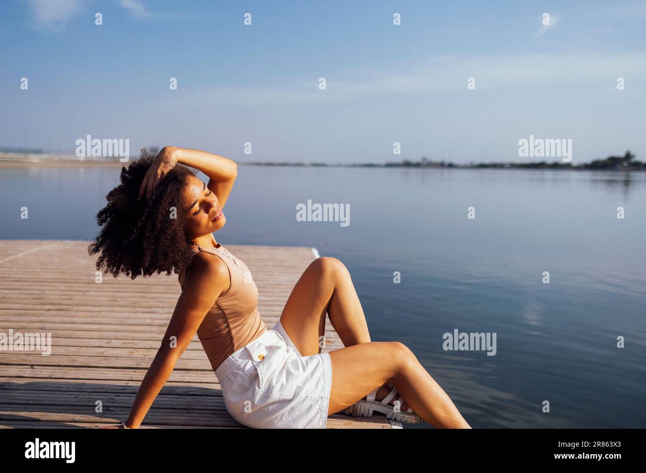 Beautiful mixed race girl is sitting on wooden pier and enjoying nature. Charming female student in beige top and white shorts has closed her eyes and Stock Photo