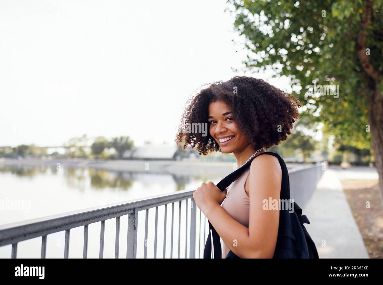Attractive mixed race girl holds backpack and smiles at camera. Happy cute african schoolgirl girl with curly hair at fence on river bank. Beautiful f Stock Photo