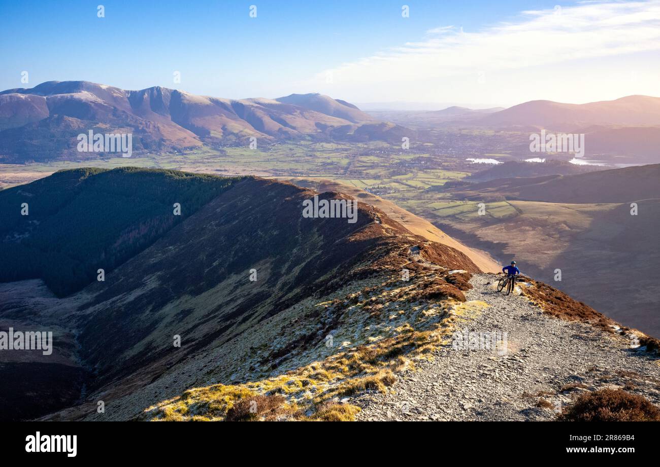 A mountain biker looking up the trail to the summit of Grisedale Pike with Skiddaw and Blencathra in the distance in winter in the English Lake Distri Stock Photo