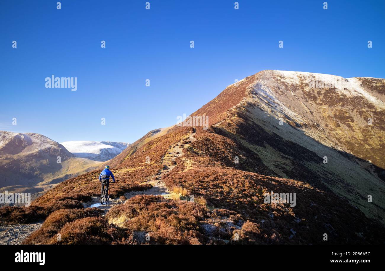 A mountain biker riding up the trail to the summit of Grisedale Pike with Crag Hill in the distance in winter in the English Lake District, UK. Stock Photo