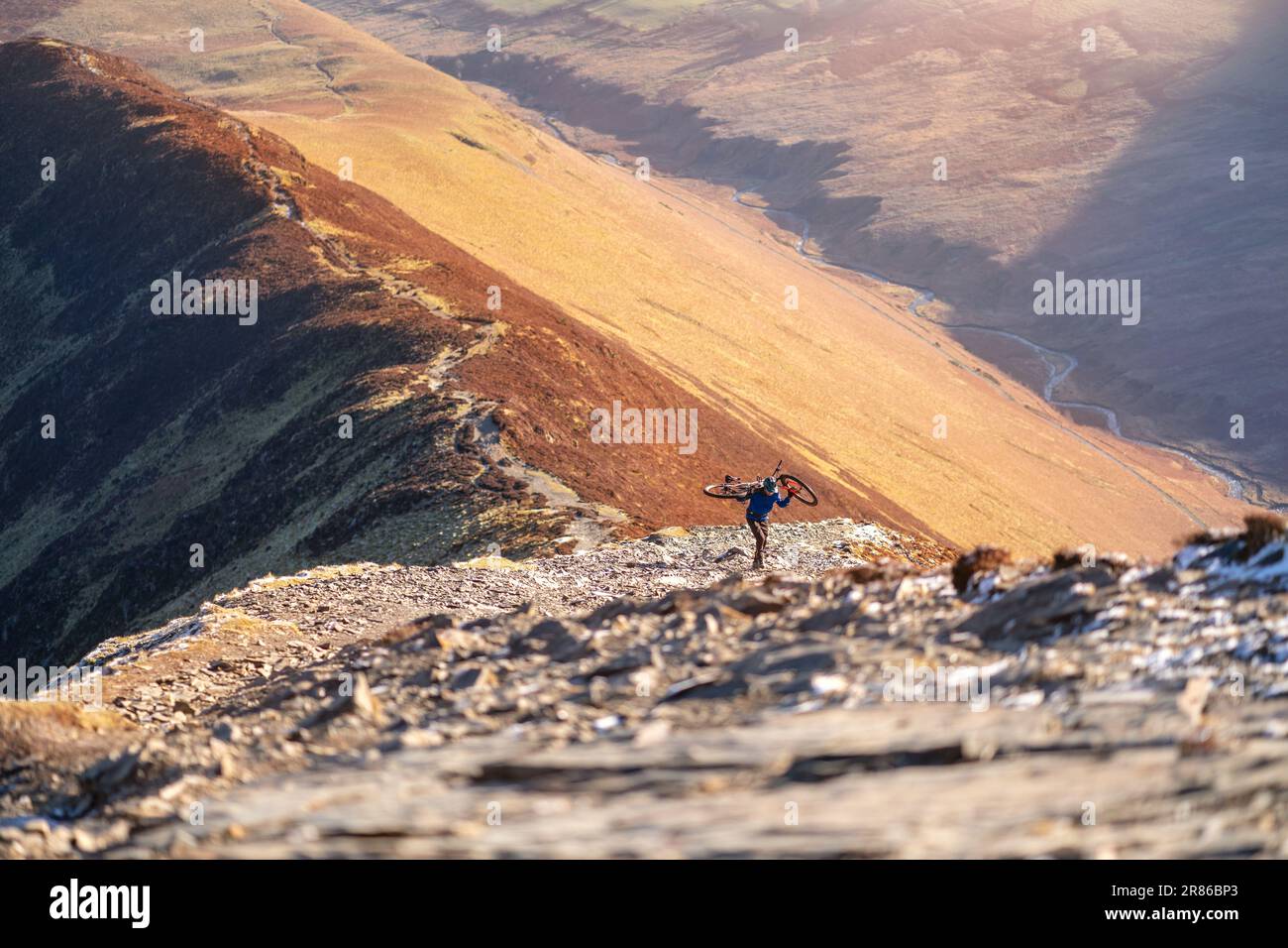 A mountain biker carrying their bike up the rocky trail to the summit of Grisedale Pike in winter in the English Lake District, UK. Stock Photo