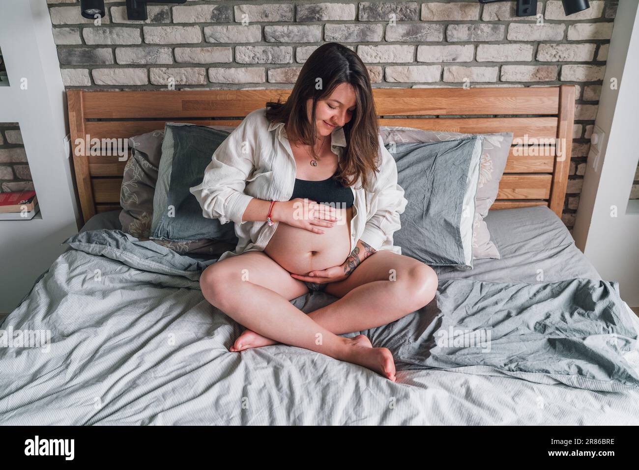 Young pregnant woman sitting with cross-legged tender touching belly on the bed in bedroom in the early morning time. Women's health, happy pregnancy Stock Photo