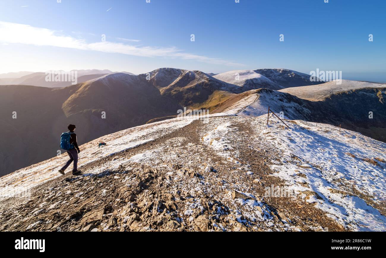 A female hiker and their dog walking over the summit of Grisedale Pike with Crag Hill and Grasmoor in the distance in winter in the English Lake Distr Stock Photo