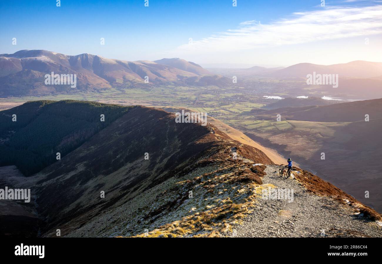 A mountain biker taking in the views on the trail up to the summit of Grisedale Pike with Skiddaw and Blencathra in the distance in winter in the Engl Stock Photo